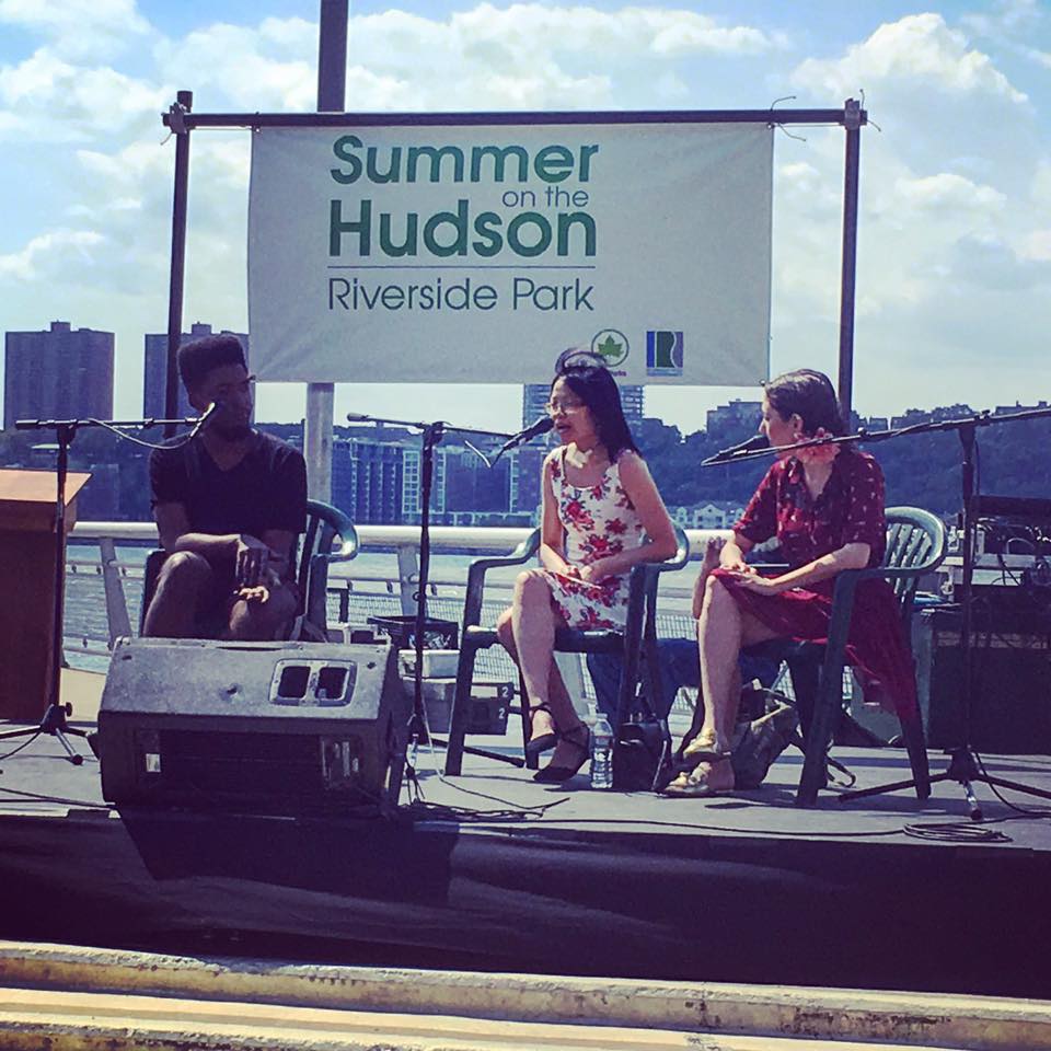Wendy is wearing a floral dress and sitting on a stage next to two other panelists on either side. They're speaking at an event called AmpLit Fest. A sign hangs above their heads with the words: "Summer on the Hudson / Riverside Park."