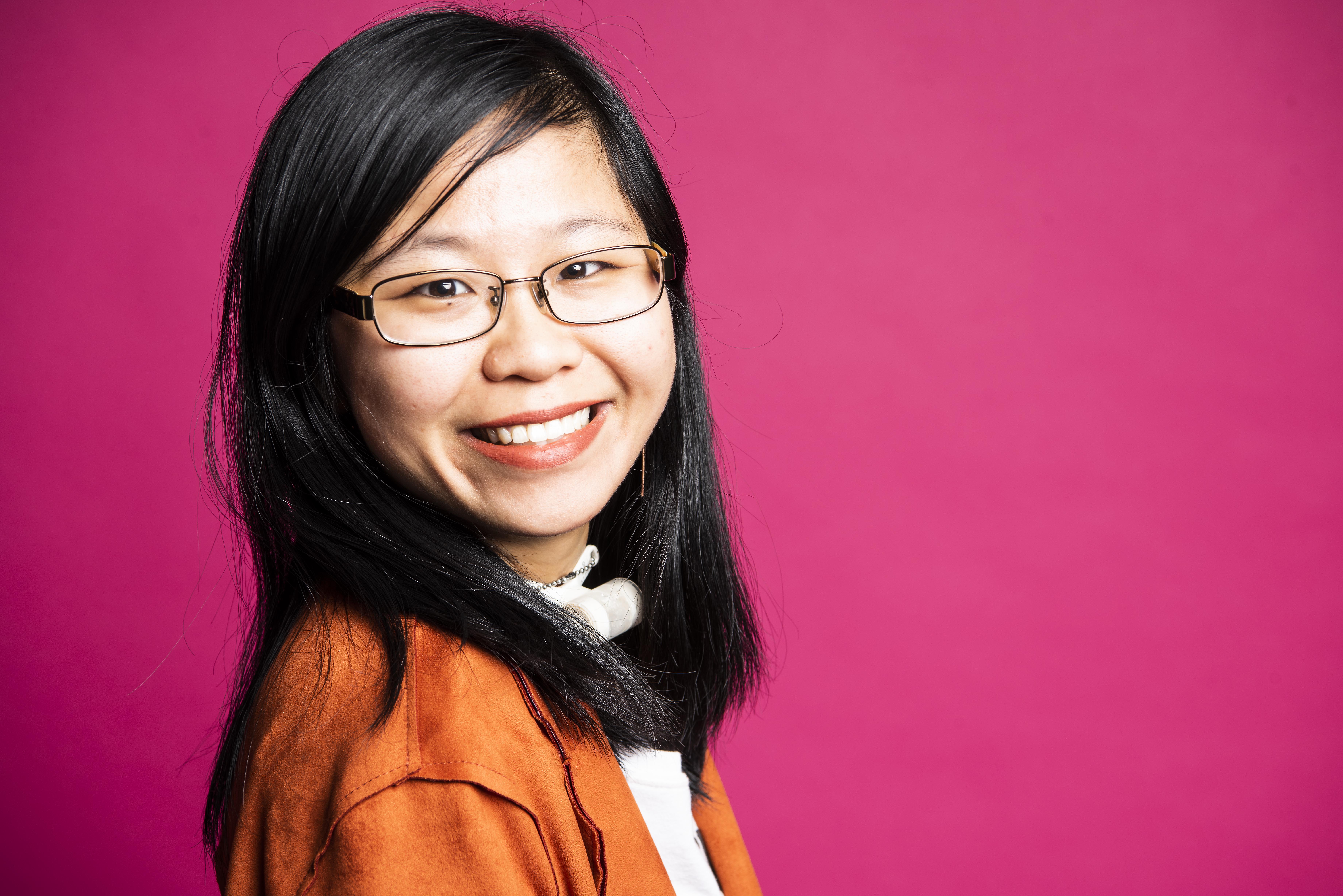 A young Chinese American woman smiles at the camera against a pink background. She's wearing glasses, a tracheostomy tube and an orange jacket.