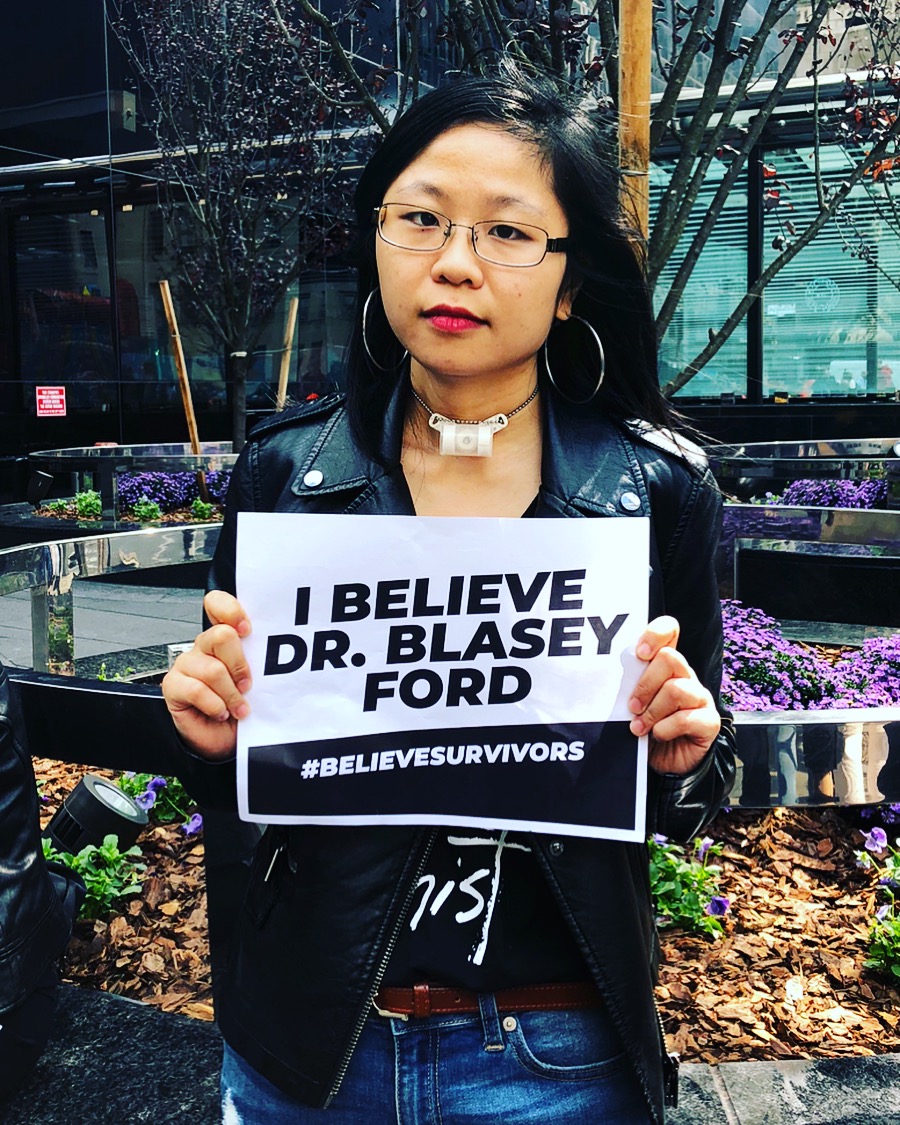 A young Chinese woman with a trach tube is wearing a black moto jacket, red lipstick, and silver hoop earrings. She's holding a piece of paper up that says: "I BELIEVE DR. BLASEY FORD" with the hashtag #BELIEVESURVIVORS at the bottom. She's outside, standing in front of a glass window.