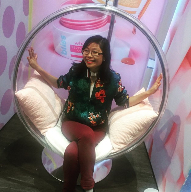 A Chinese woman with black medium-length hair sits on a plastic round bubble chair with pillows. She's wearing a flower bomber jacket and burgundy pants. The background has pink polka dots and shows a giant picture of Bliss skin care.