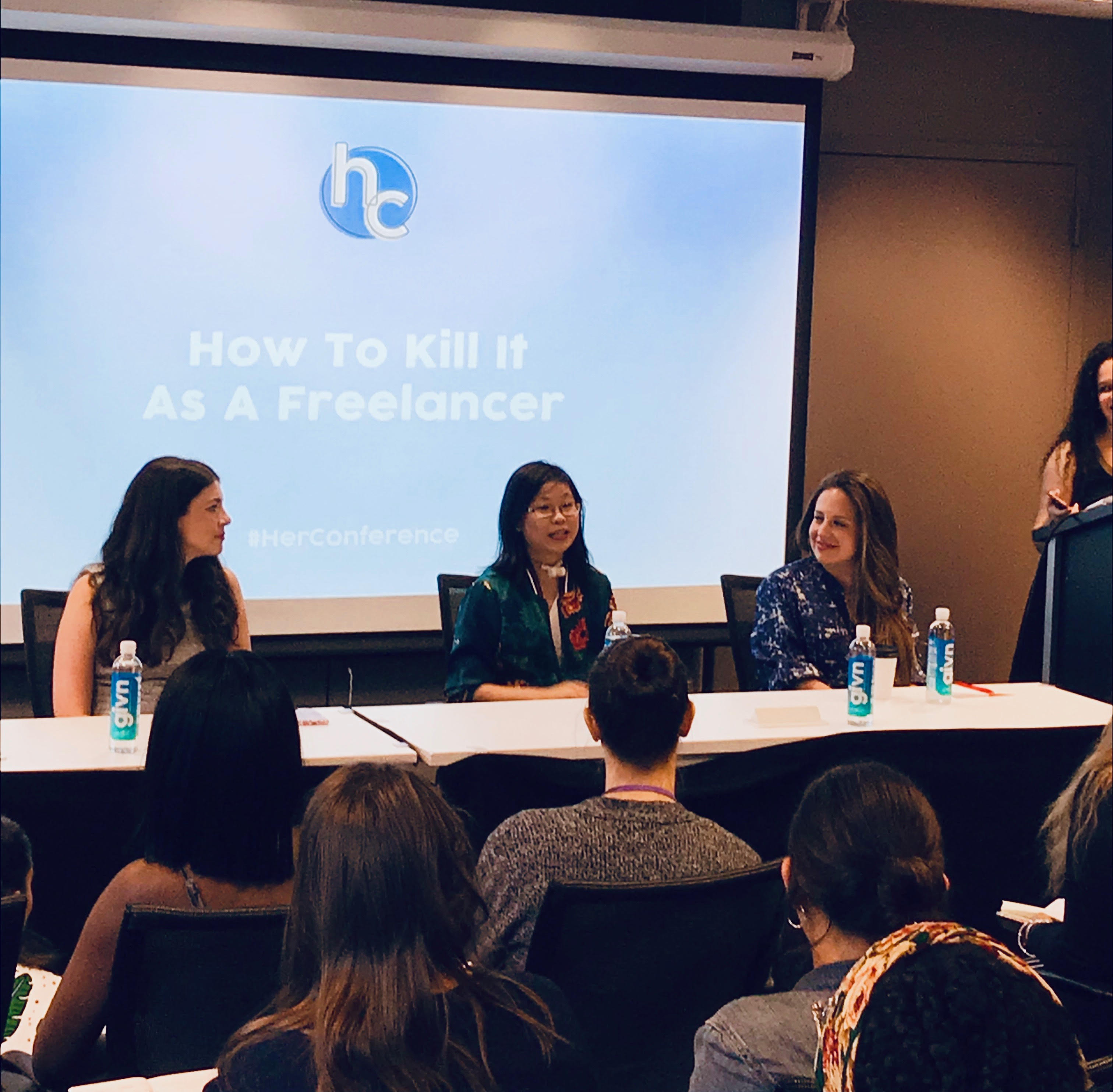 A Chinese woman with a trach tube is speaking on a panel with four other women at the HerCampus Conference in 2018. She's in front of a presentation titled, "How To Kill It As A Freelancer." Several audience members are sitting in the foreground.